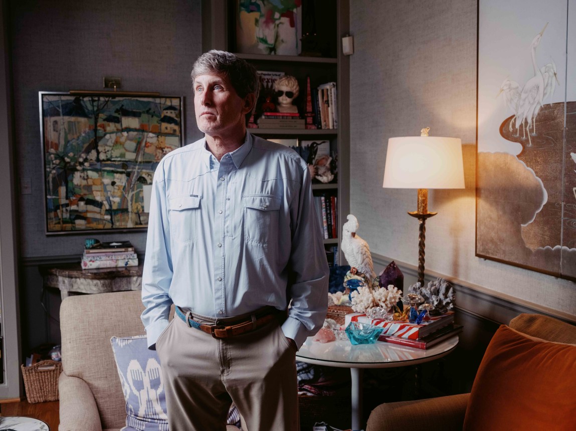 A man with gray hair, wearing a light-blue button-down shirt and tan trousers, stands in a room with shelves and artwork behind him. He has his hands in his pockets and is looking to his right.