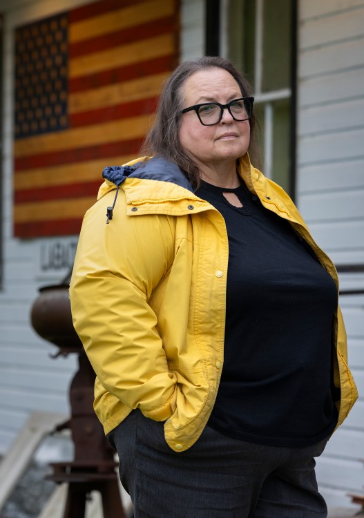 A woman wearing black frame glasses, a yellow rain jacket over a black shirt and black jeans poses for a portrait with her hands in her pocket, in front of a house with white siding and an American flag.