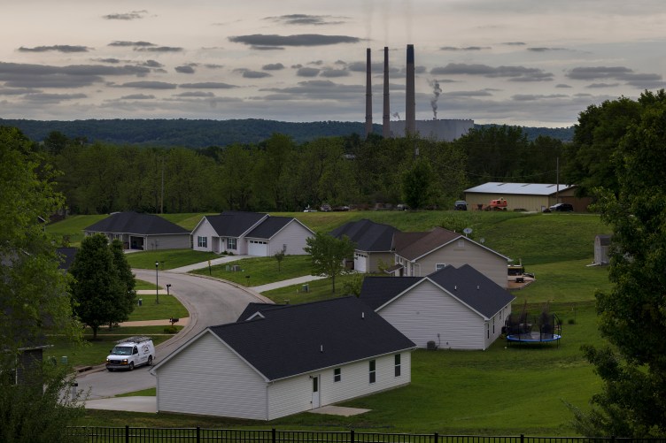 Thin plumes of smoke rise from three slender, tall smoke stacks, behind a residential neighborhood with large manicured grass yards surrounded by forests.