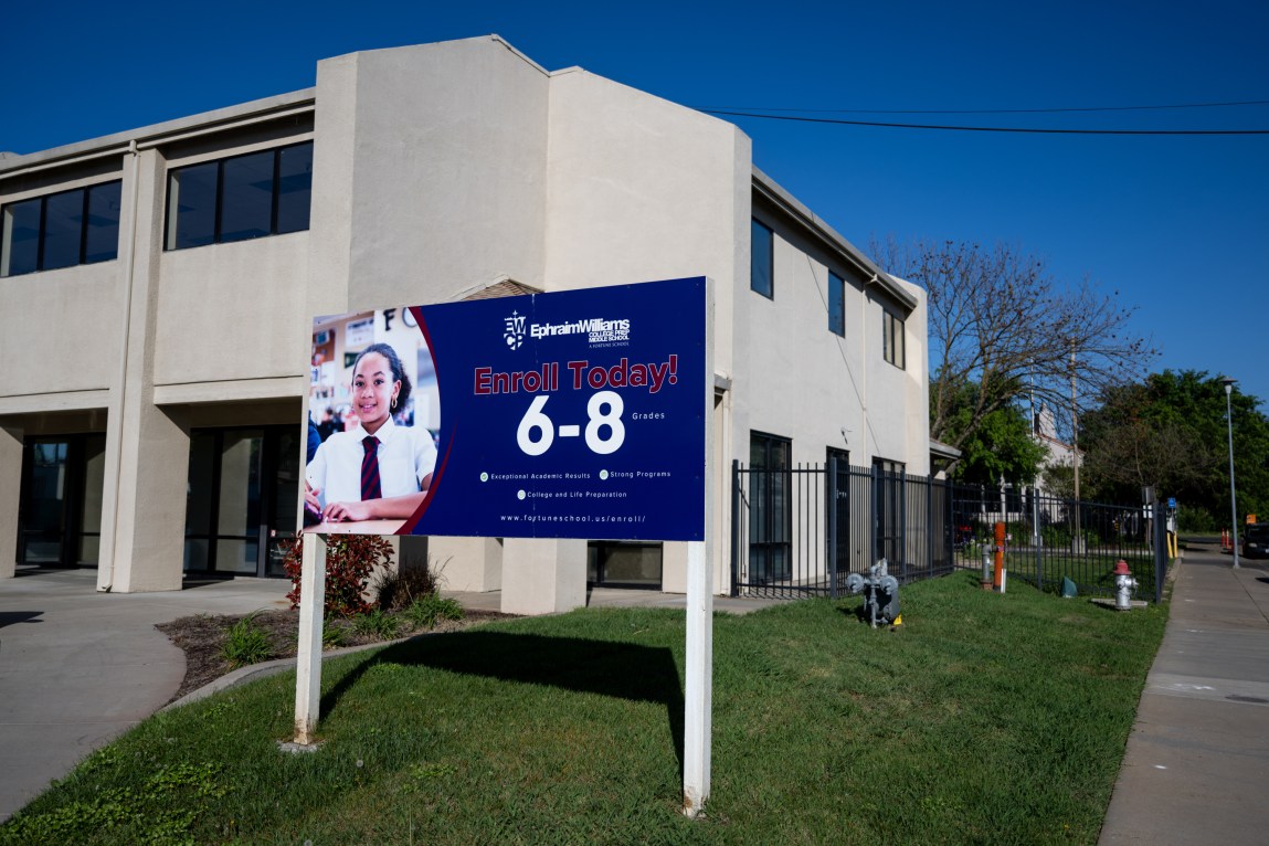 A school building with a sign in front of it that shows a photograph of a student and text that reads, “Enroll Today! 6-8 grades.”