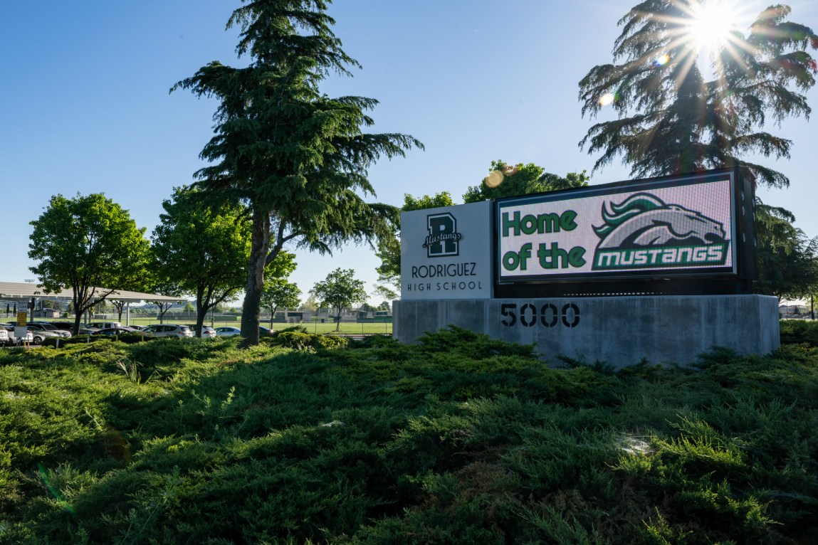A sign that reads, “Rodriguez High School,” and, “Home of the Mustangs,” outside surrounded by trees and bushes.