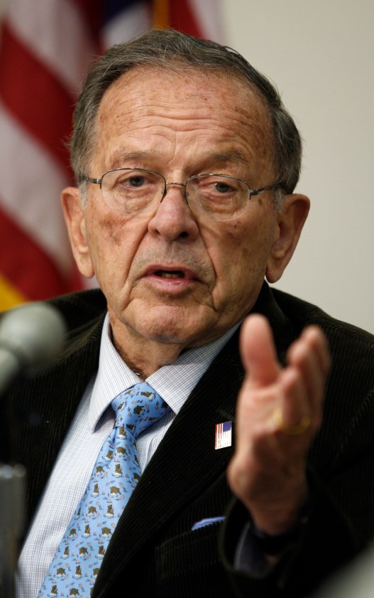 A man with gray hair and glasses wearing a tie and U.S. flag lapel pin gestures with his hand toward the camera. An out-of-focus U.S. flag can be seen behind him.
