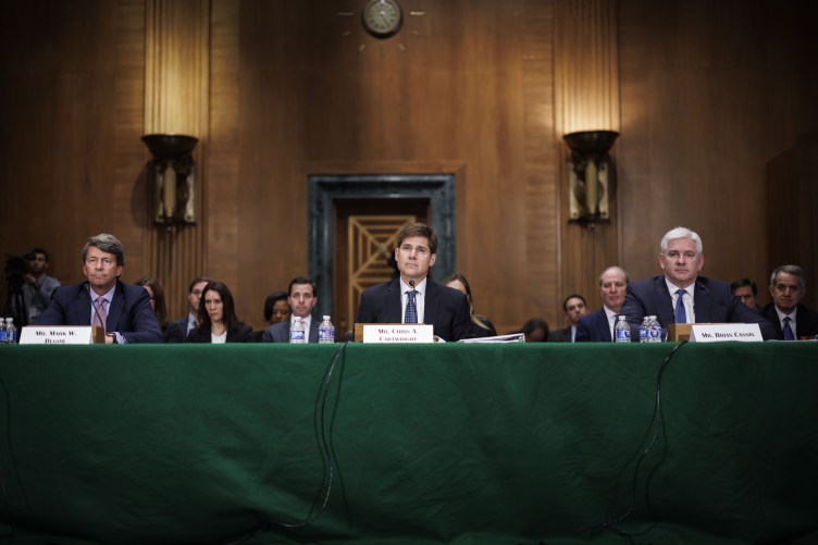 Trois hommes en costume sont assis à une table verte, derrière eux des gens sont assis dans une pièce lambrissée.