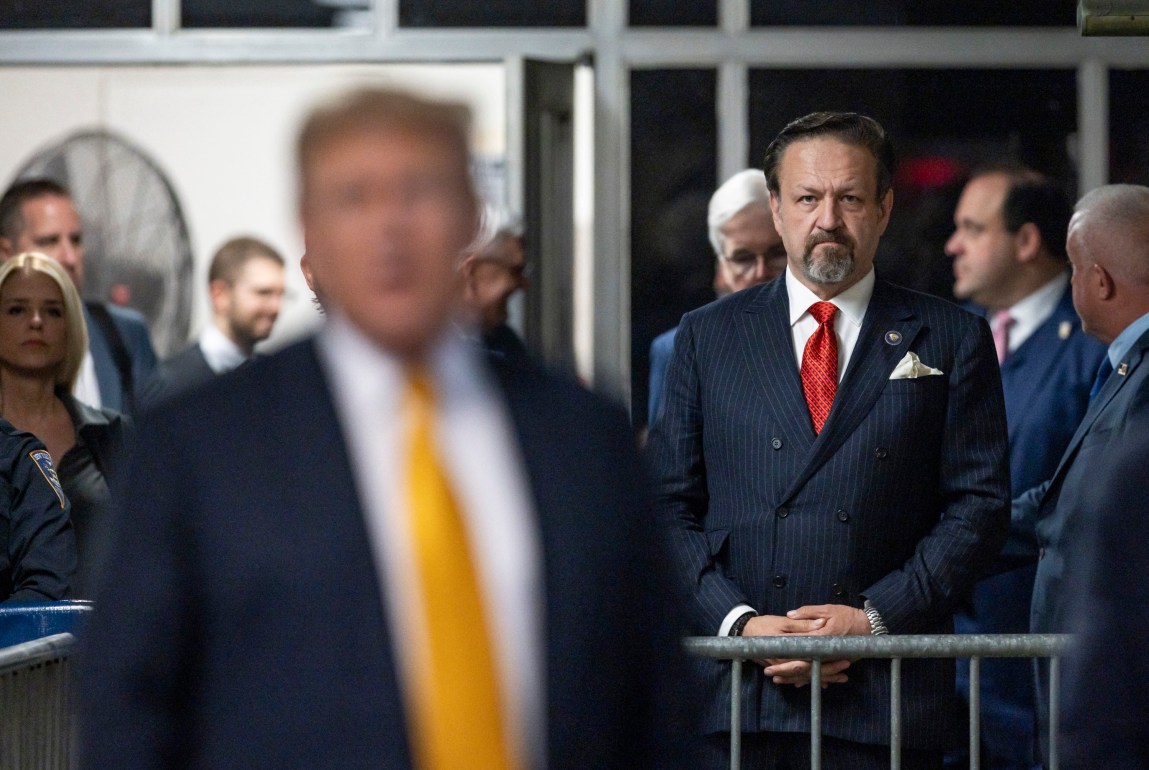 A man in a suit with a serious expression stands behind a gate with his hands clasped together. He has his eyes fixed on the foreground, where President Donald Trump is a blurred figure addressing a crowd.