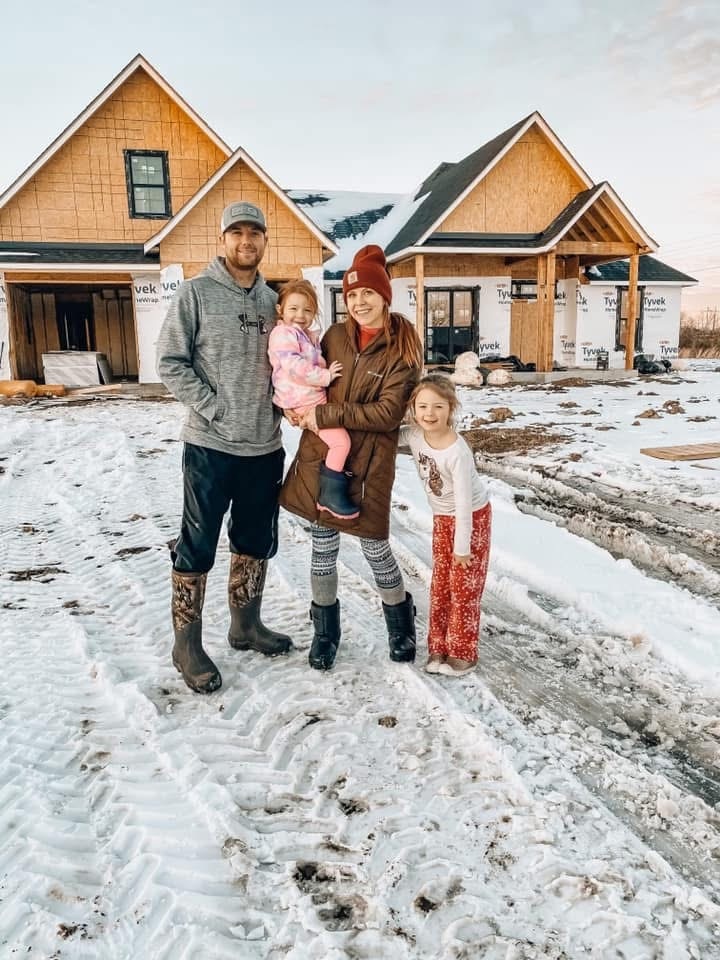 A man, a woman holding a young girl, and another girl stand together in the snow, posing for a photo in front of a large house that is being built.