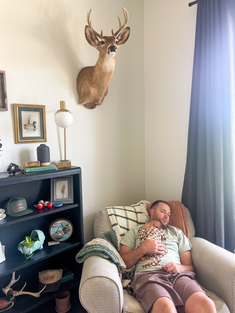 A man sitting in a large chair sleeps with a baby boy on his chest. A deer head is mounted on the wall above him.
