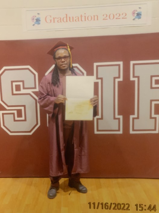 A man in a red graduation gown and cap holds a diploma up for the camera under a sign that reads, “Graduation 2022.”