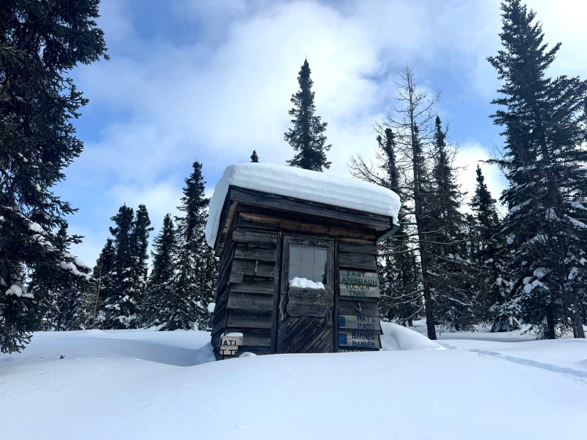 Coniferous trees surround a lone wooden cabin sitting on a snowy hill with snow covering the roof. In the background is a blue sky with white clouds.