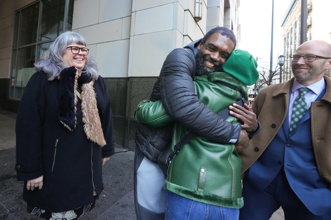 A man and woman hug as another woman and man, smiling, look on in front of a courthouse.