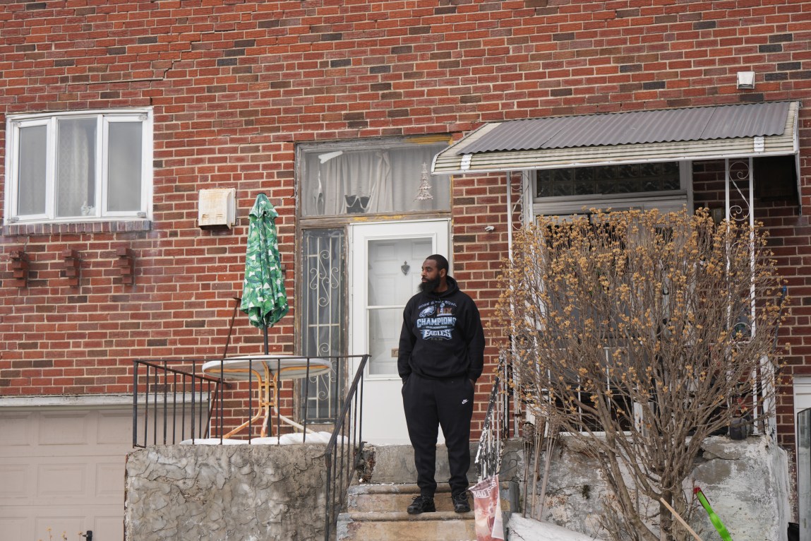 A man stands on the steps in front of a brick building. Next to him is a patio with a table and umbrella and the front entrance to a home.