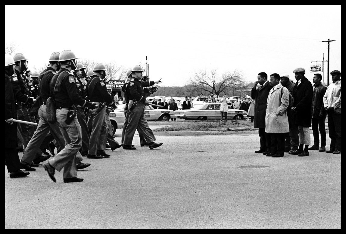 Une photo en noir et blanc montre une file de policiers avançant depuis la gauche, matraques dégainées, et un groupe d'hommes noirs rassemblés sur la droite. Une foule regarde en arrière-plan.