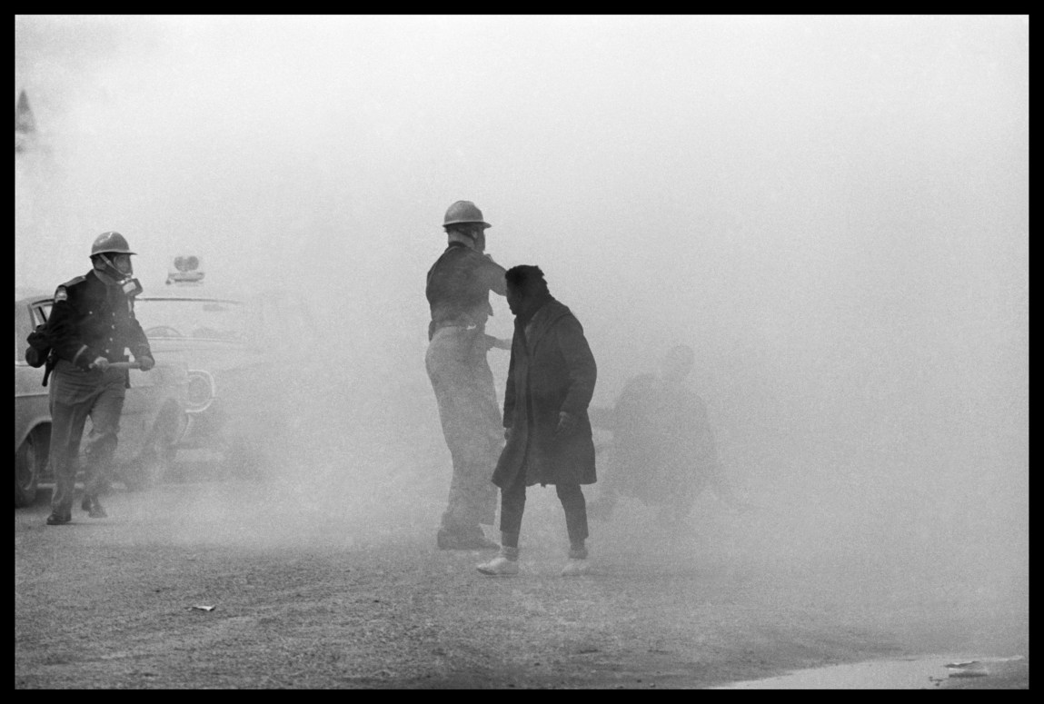 Une photo en noir et blanc d'une scène obscurcie par un nuage de gaz lacrymogène. Deux policiers et un autre homme sont visibles. Une autre personne est à peine visible dans la brume alors qu’elle tombe au sol.