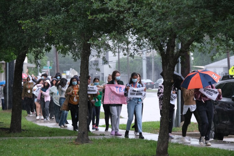 Eine Gruppe von Studenten marschiert während einer Protestaktion mit Regenschirmen und Schildern über einen verregneten, von Bäumen gesäumten Gehweg.