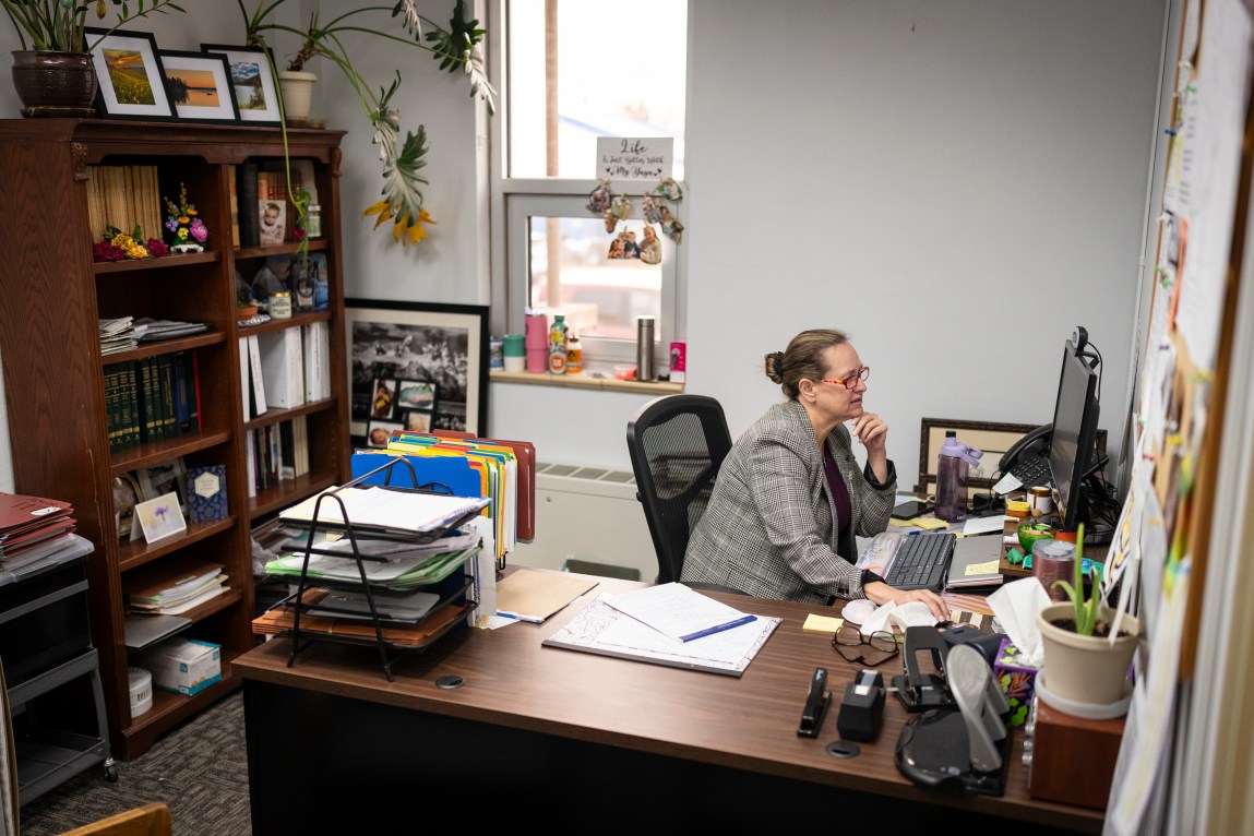 A woman sitting at an office desk working on a computer. The office includes a large wooden bookshelf filled with books and binders, various desk organizers, files and personal photos.