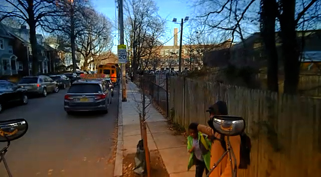 A camera view from the exterior of a school bus shows a boy and a man in front of the bus as it moves onto a city sidewalk.
