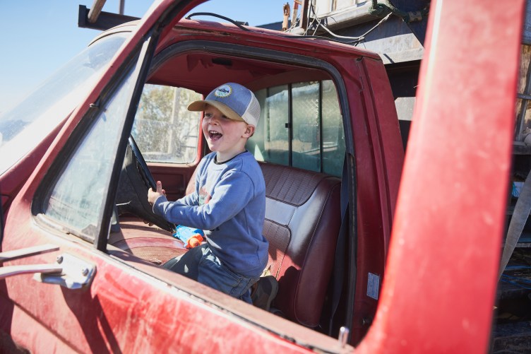 A young boy sits in the driver’s seat of a red truck, holding the steering wheel and smiling. He is framed by the open car door.