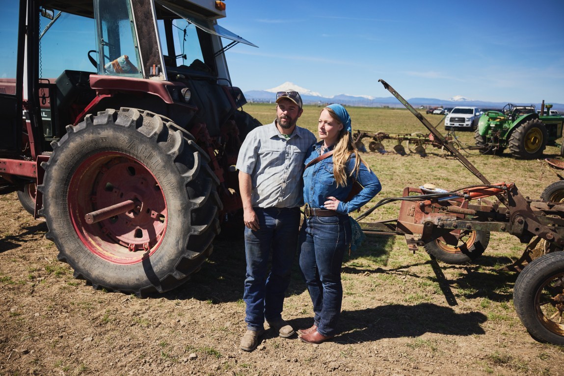 A man and a woman stand together in front of a tractor. Behind them is another tractor, cars, a wide-open landscape and, in the distance, snow-capped mountains.