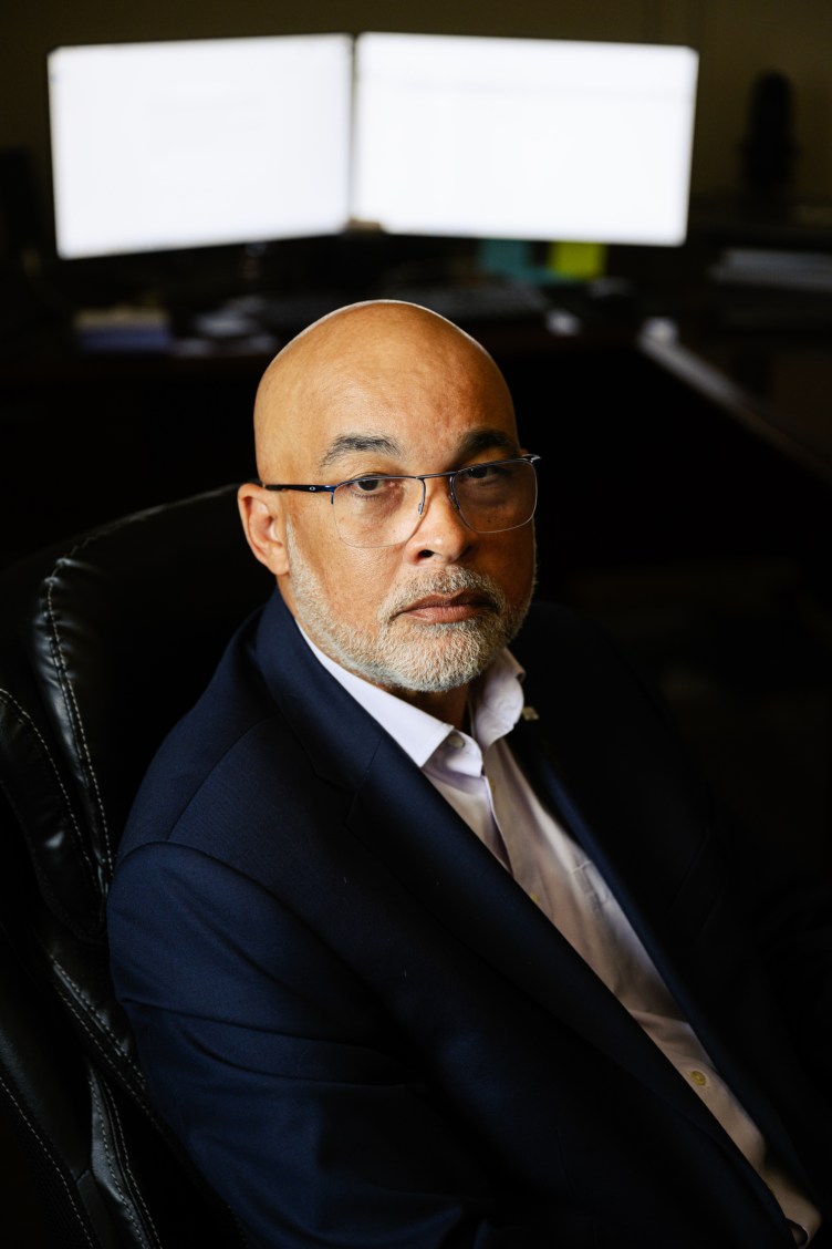 A portrait of a bald man with a gray beard and glasses, wearing a navy blue blazer and a white button-down shirt. He is sitting in a black leather office chair with a serious expression, and two illuminated computer monitors behind him.