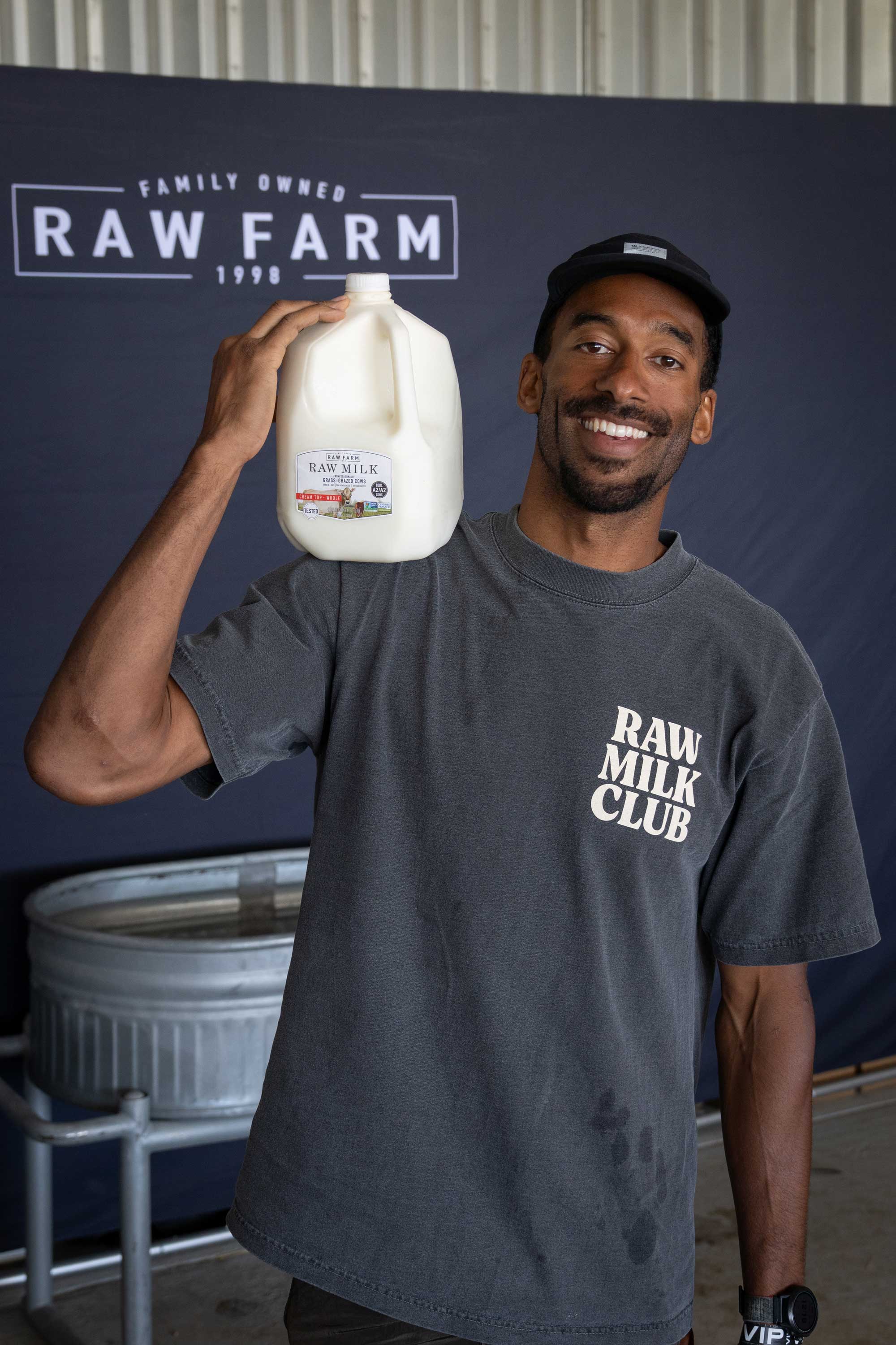 A smiling man wearing a black cap and a “Raw Milk Club” T-shirt holds a gallon jug of milk on his shoulder, standing in front of a blue Raw Farm backdrop.