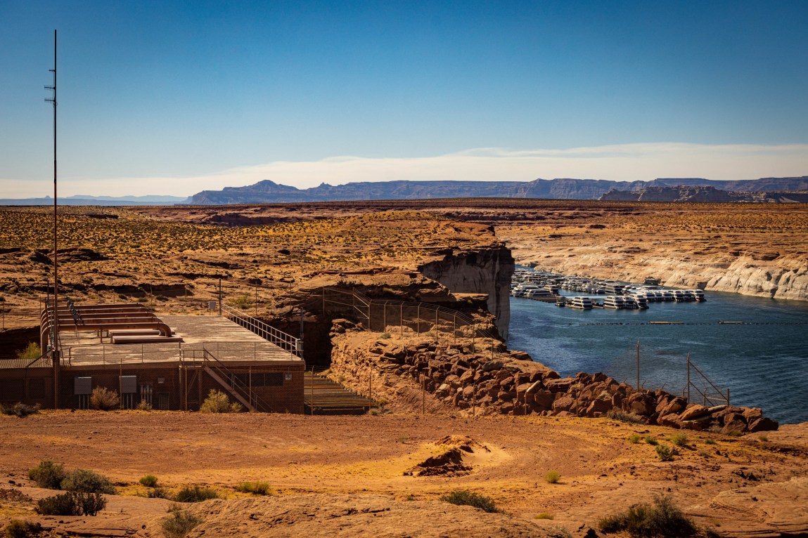 An industrial water pumping station with a tall antenna sits on a rocky bluff overlooking a large river canyon. In the distance, numerous white houseboats are moored together along the water.