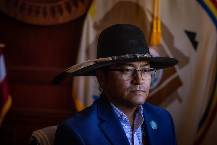 A portrait of a man wearing a blue suit jacket, a striped collared shirt and glasses. He wears a wide-brimmed black hat adorned with silver accents and a feather, and is looking slightly off-camera against a background featuring a tribal flag.