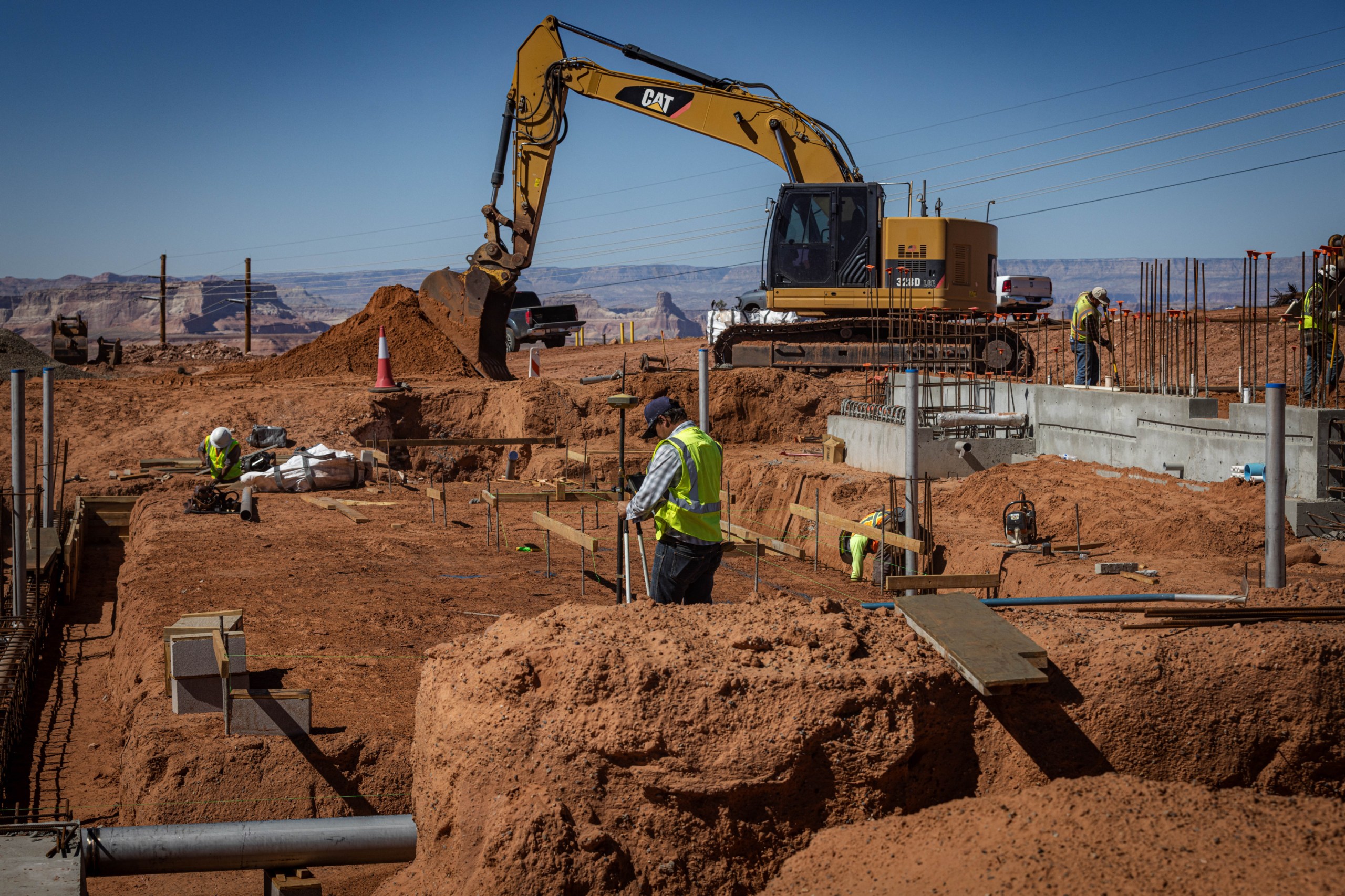 Construction workers in high-visibility vests work at a red-dirt excavation site alongside concrete foundations, while a large yellow excavator digs in the background.