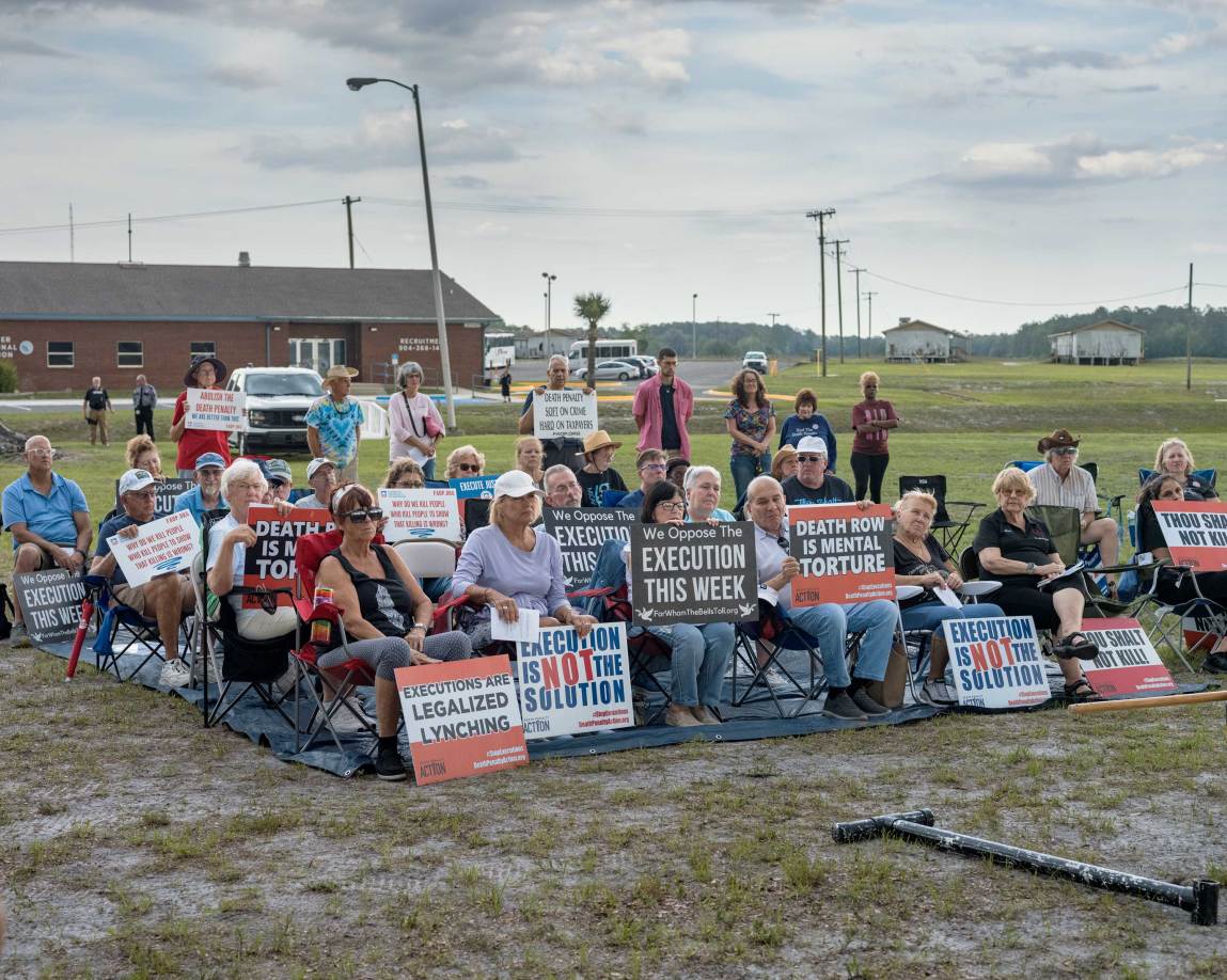 A wide shot shows a group of anti-death-penalty protesters seated closely in lawn chairs arranged on a large dark tarp outdoors. They are holding various signs with messages such as “We Oppose the Execution This Week,” “Death Row Is Mental Torture,” “Execution Is NOT the Solution,” and “Executions Are Legalized Lynching.”