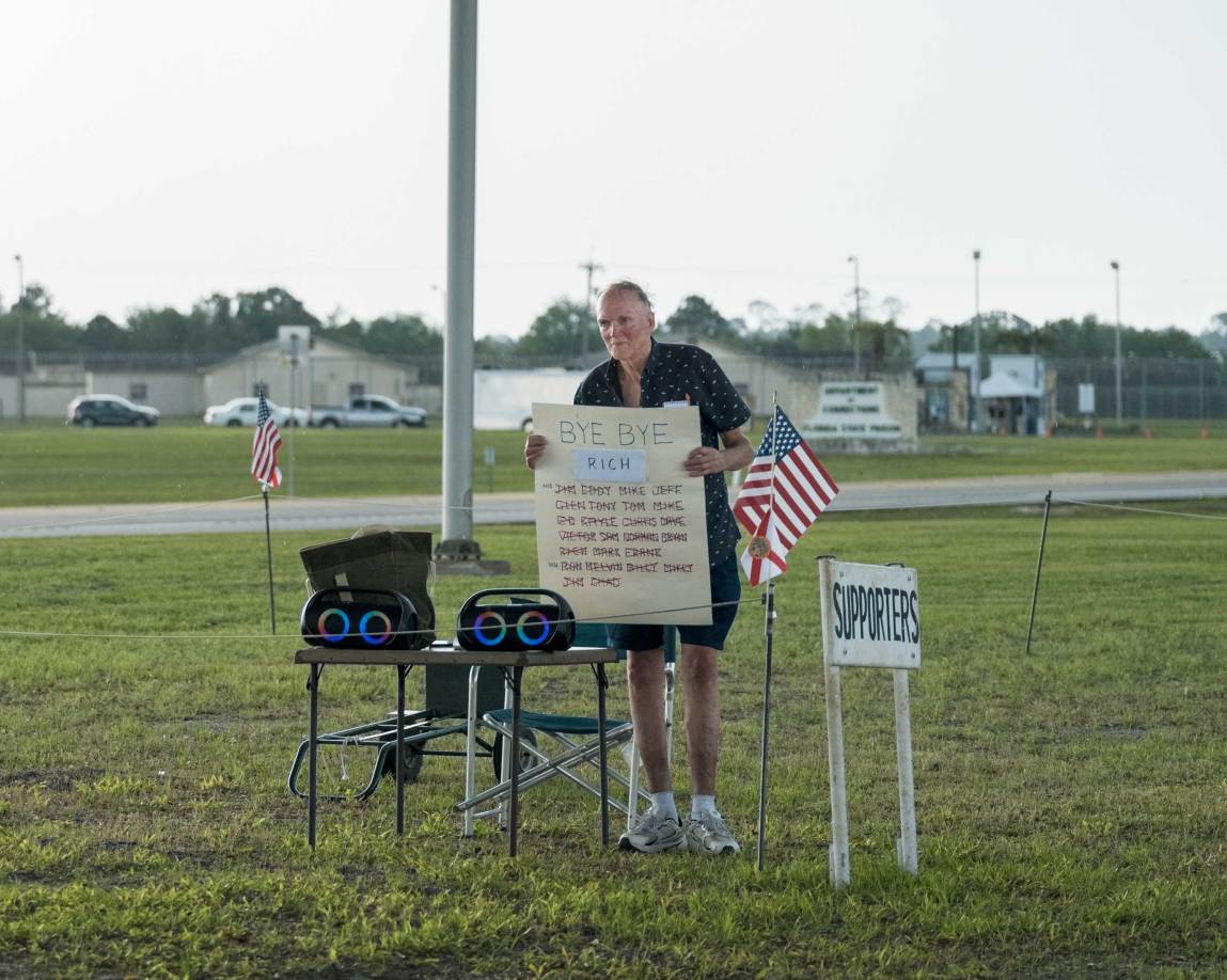 A man stands in a grassy field holding up a large sign that reads “"Bye Bye Rich” at the top, followed by rows of handwritten first names. He is standing behind a small folding table equipped with two speakers displaying glowing blue and green lights.