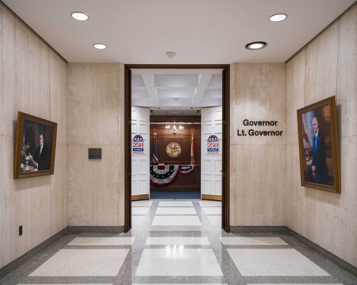 A symmetrical view down a marble-tiled hallway leads to a set of open double doors labeled "Governor/Lt. Governor" on the right wall. Inside the wood-paneled office beyond the doors, an American flag and a Florida state flag stand on either side of a large gold seal mounted on the wall.