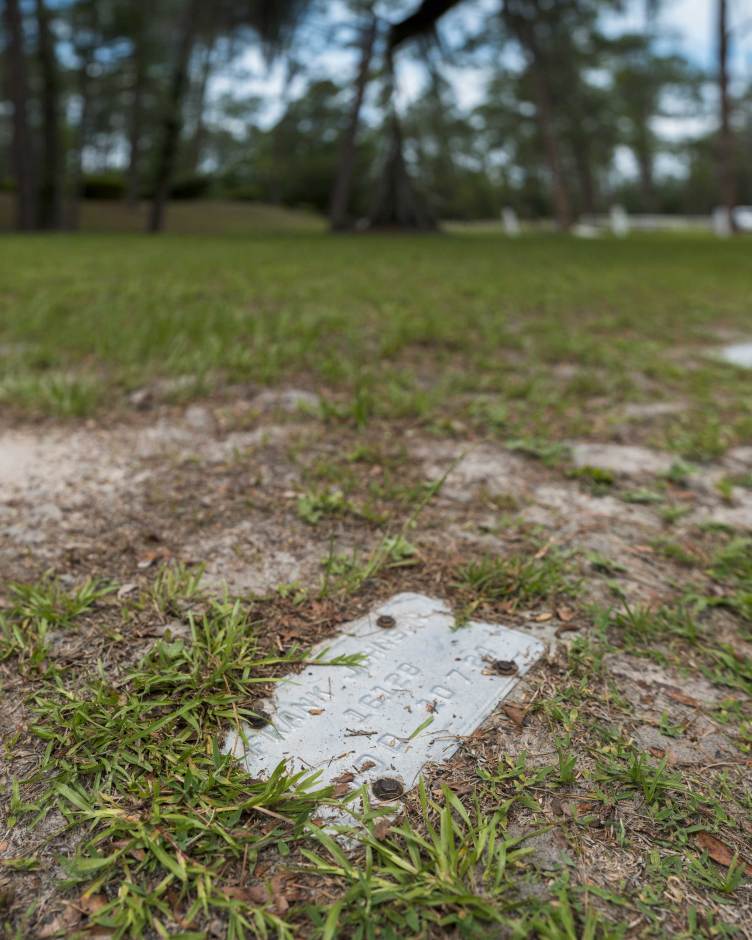 A low-angle shot focuses on a weathered, rectangular metal grave marker embedded flush with the sandy soil and patches of green grass.