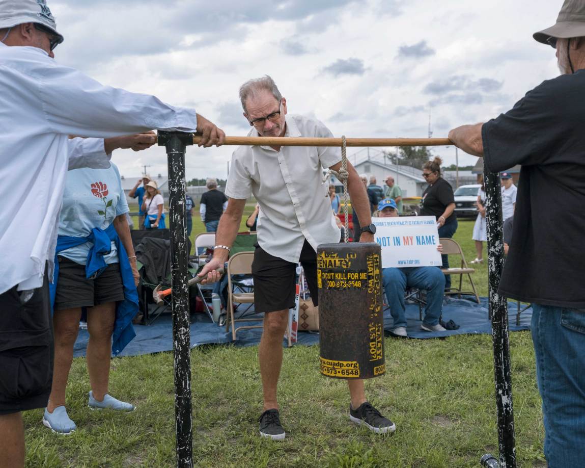 A medium shot captures a man with glasses and a white short-sleeved button-down shirt standing in a grassy field, preparing to strike a large, heavy metal cylinder suspended by a rope from a horizontal wooden beam.