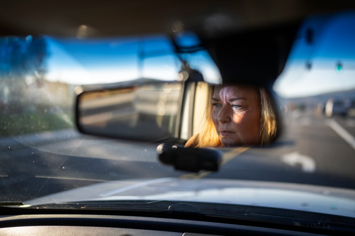 A close-up shot looking through a car's windshield, capturing a woman's reflection in the rearview mirror. She has blonde hair and a serious expression as she drives down a road in daylight.