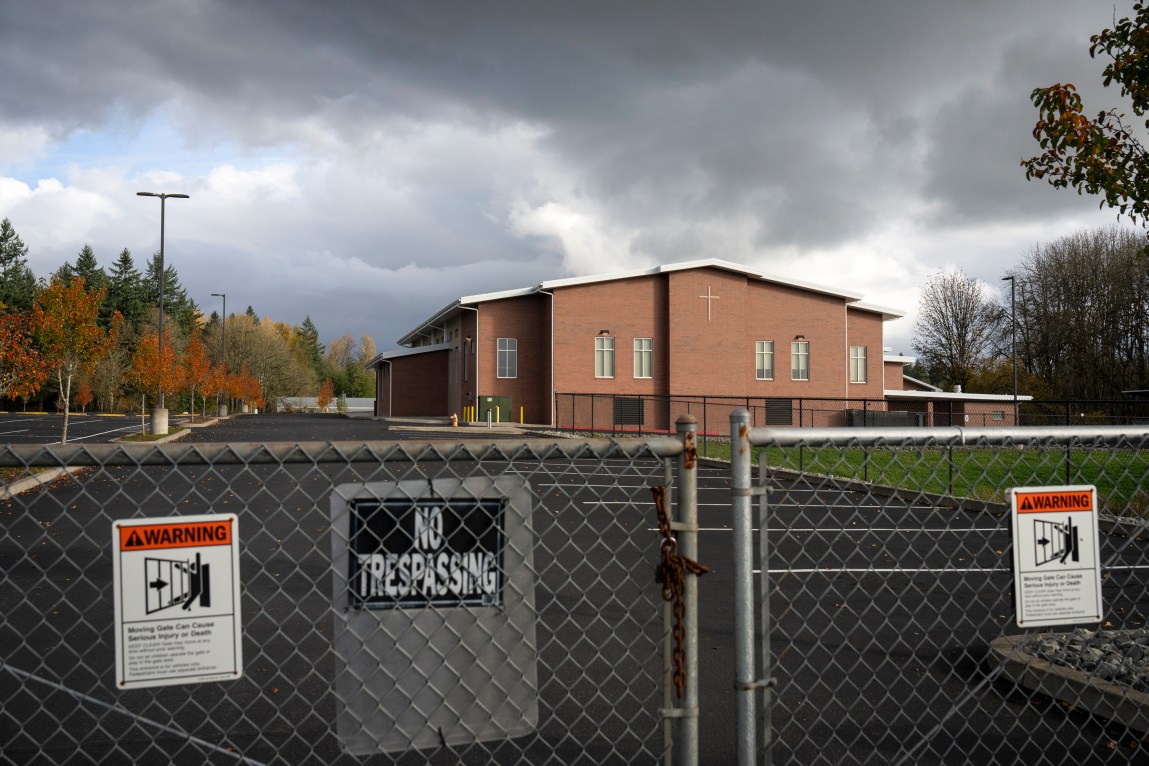 A view of a red-brick church building from behind a closed chain-link fence. The fence features a prominent "No trespassing" sign, with an empty asphalt parking lot stretching out toward the building under a cloudy sky.