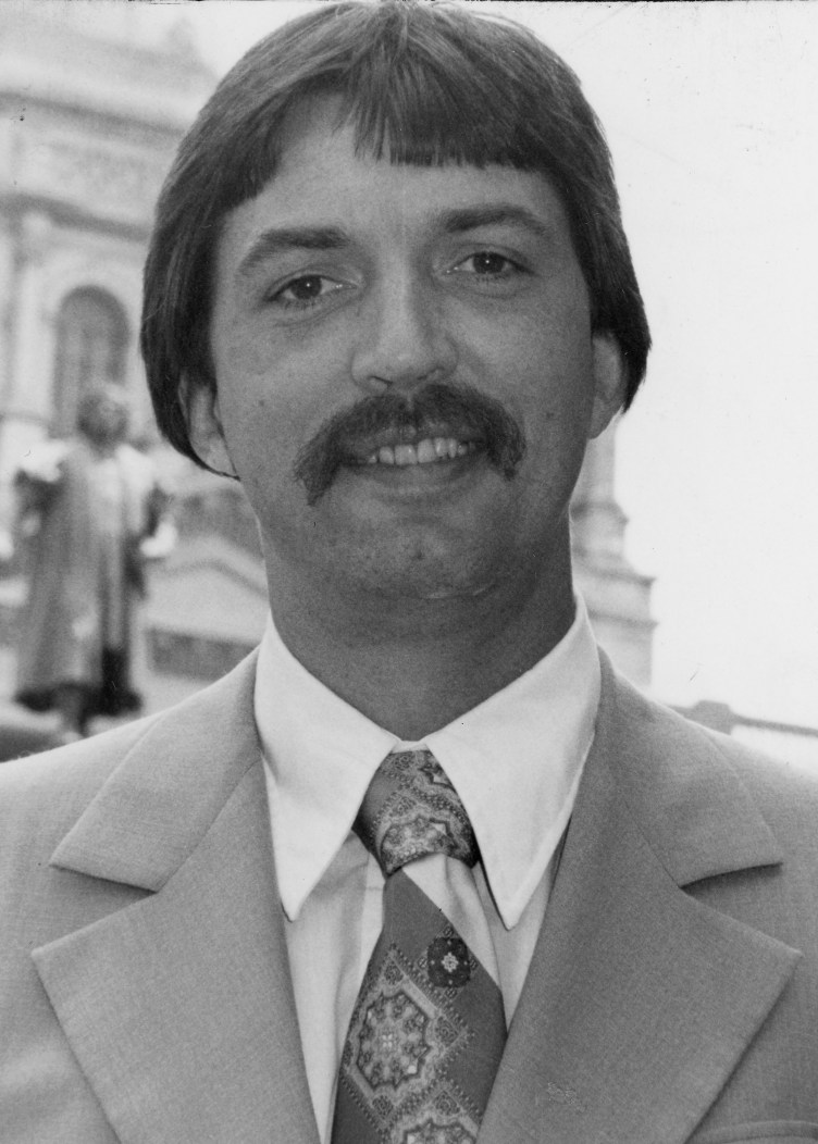 A man with a mustache, suit and paisley tie smiles at the camera.