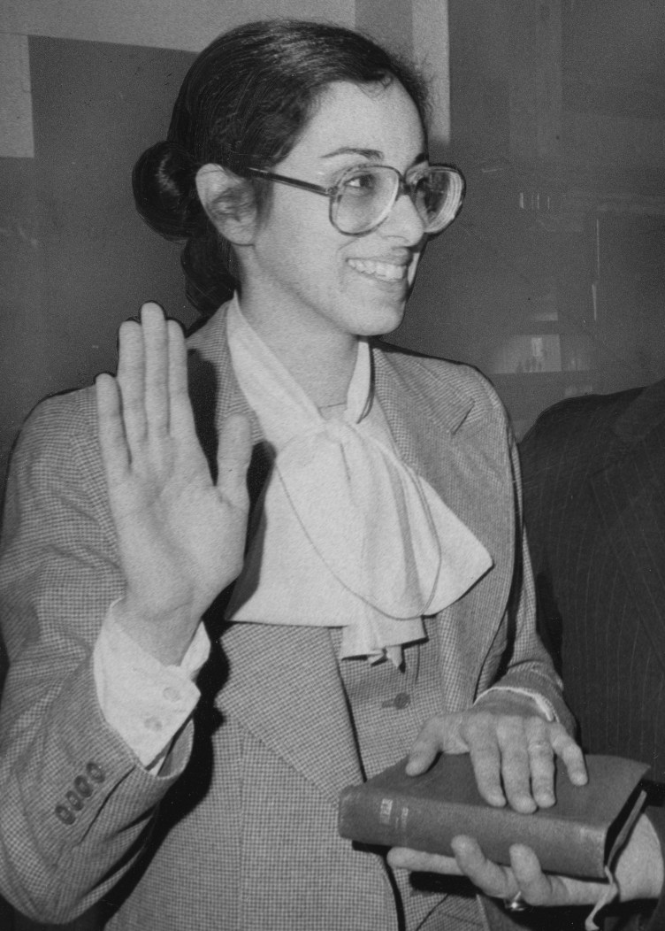 A young woman in glasses and a business suit holds her right hand up while her left rests on a book.