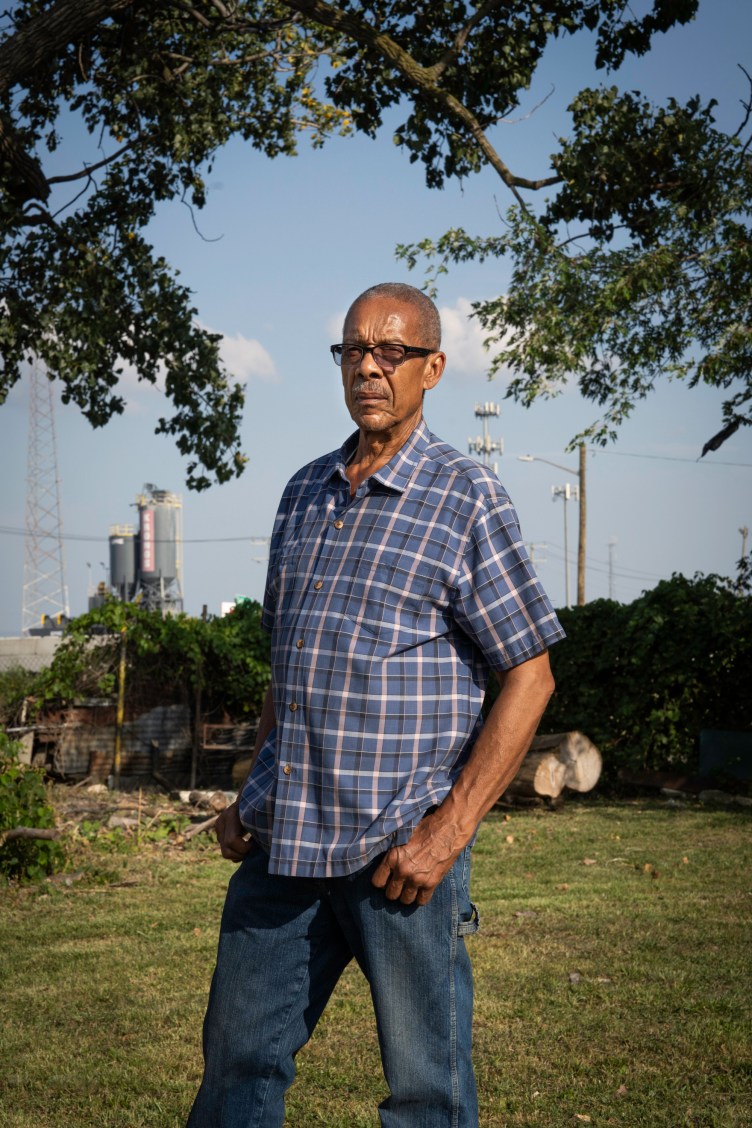 A man stands outdoors with his hands on his hips, looking at the camera with a serious expression. Behind him in the background is a large industrial structure with the word “Kronos” written on it.