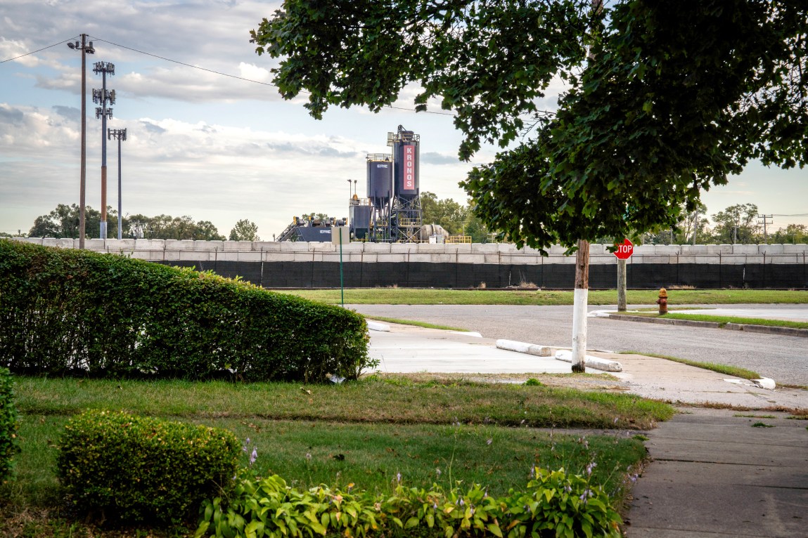 Green lawns, bushes, streets and sidewalks, with a large industrial structure with the word “Kronos” written on it in the background, overlooking the neighborhood.