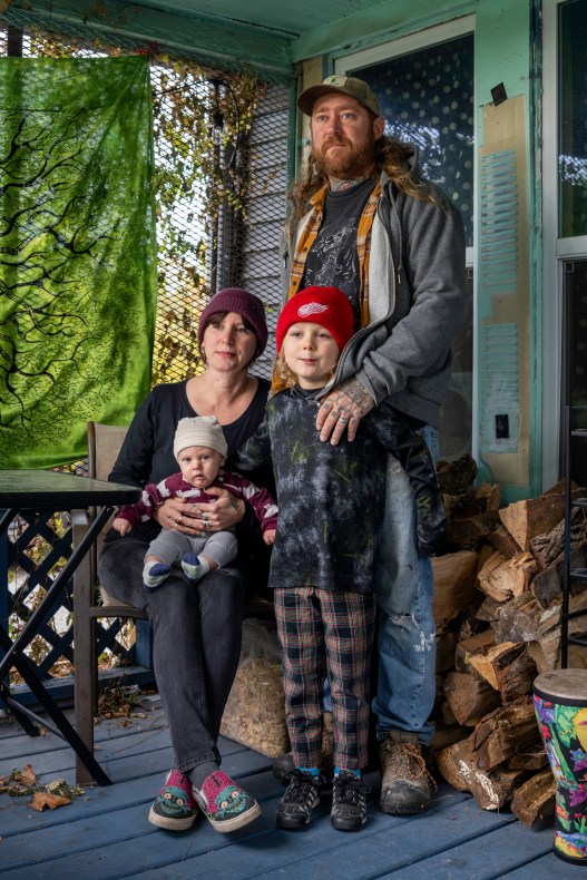 A man stands with his hand on the shoulder of a boy in front of him. Next to them, a woman sits with a baby in her lap. The four of them are on the front porch of a house.