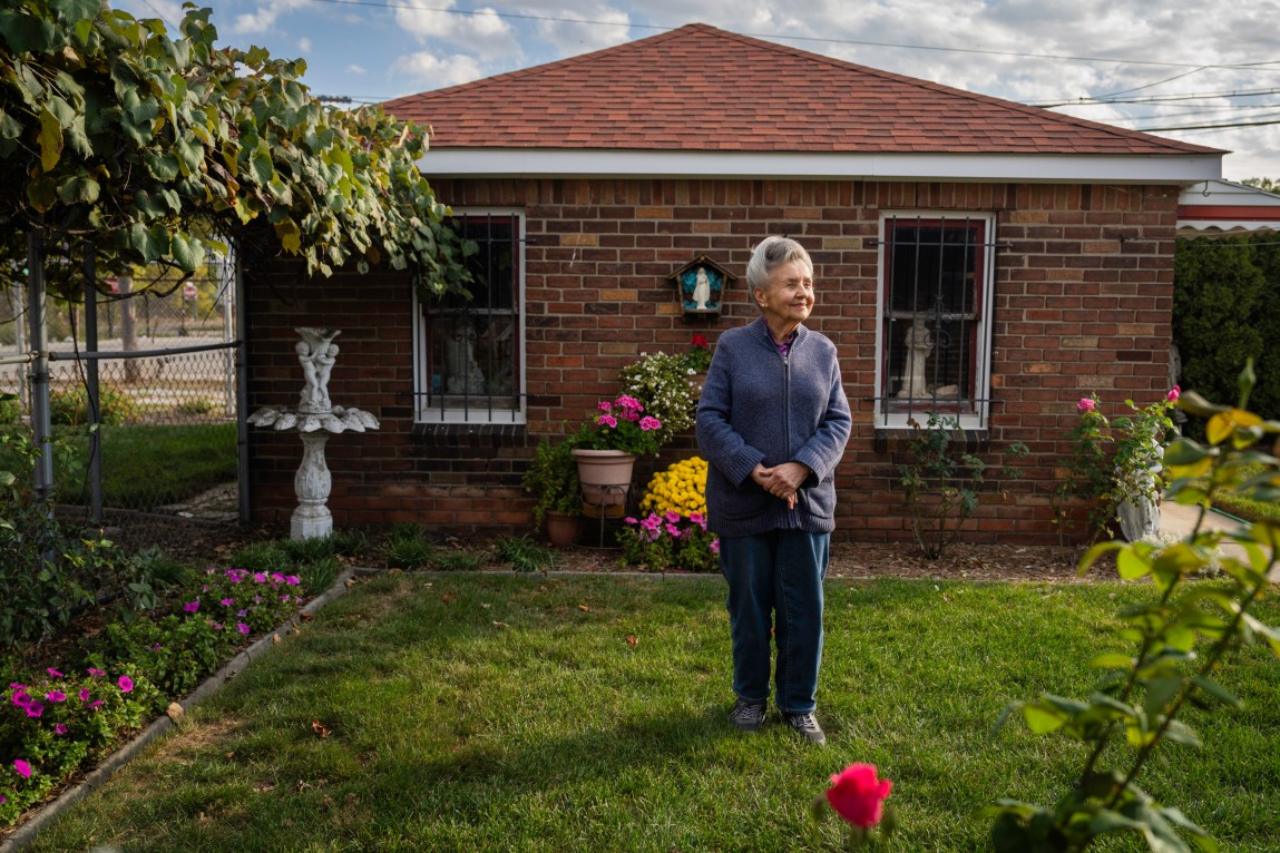 A woman stands on a lawn, smiling, in front of a small brick building. Around her, on the edge of the lawn, are small gardens with colorful flowers and small statues.