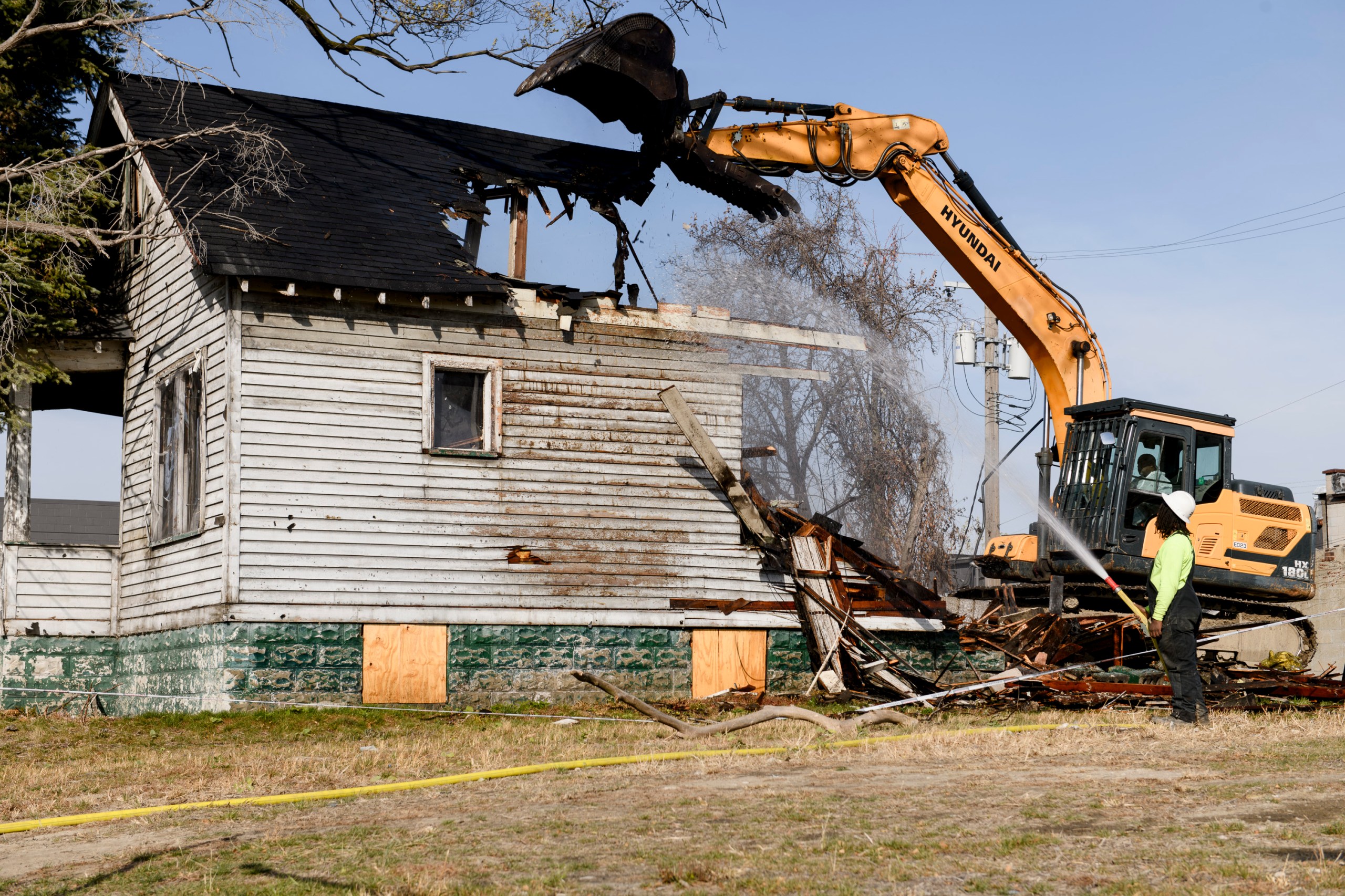 A house that is halfway through being demolished by an excavator. A person sprays water on the house with a high-powered hose.