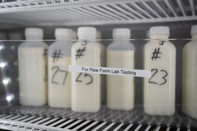 Nummerierte Milchflaschen auf einem Kühlschrankregal hinter einem Klebeband mit der Aufschrift „Für Raw Farm Lab Testing“.