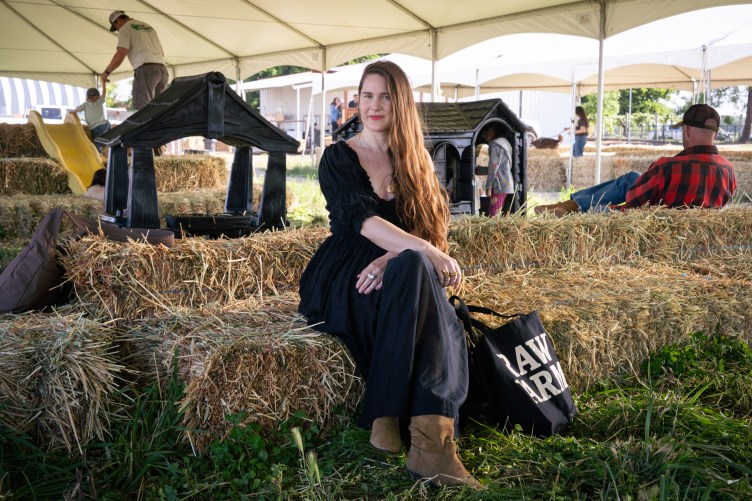 Eine Frau in einem schwarzen Kleid sitzt auf einem Heuballen neben einer Raw Farm-Einkaufstasche unter einem weißen Zelt, im Hintergrund sind andere Menschen.
