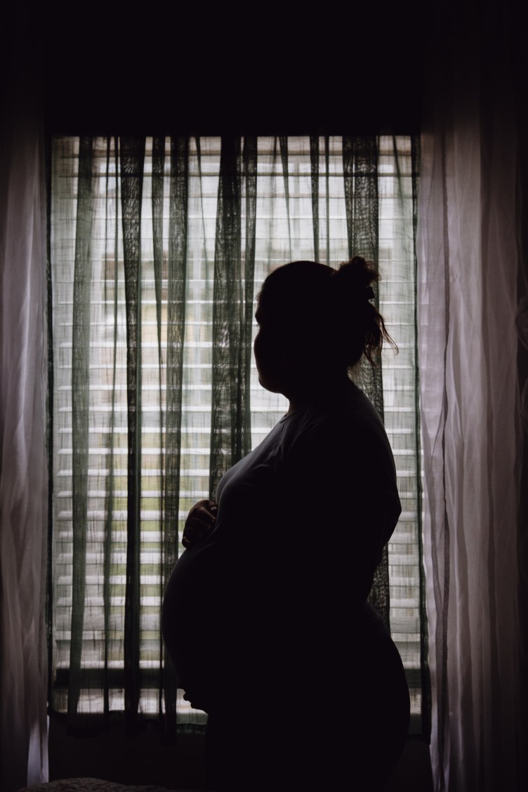 The silhouette of a pregnant woman standing in profile before a window with closed blinds and sheer curtains.
