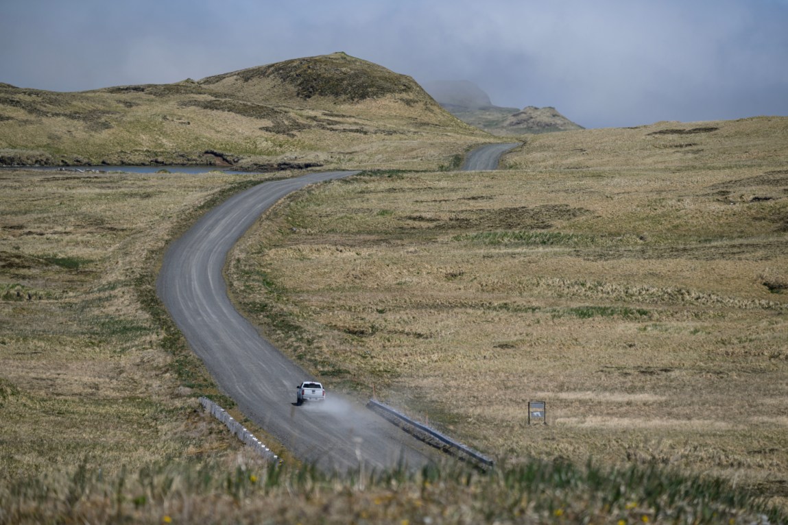 A lone white car drives along a gray road leading over a brown mountain.