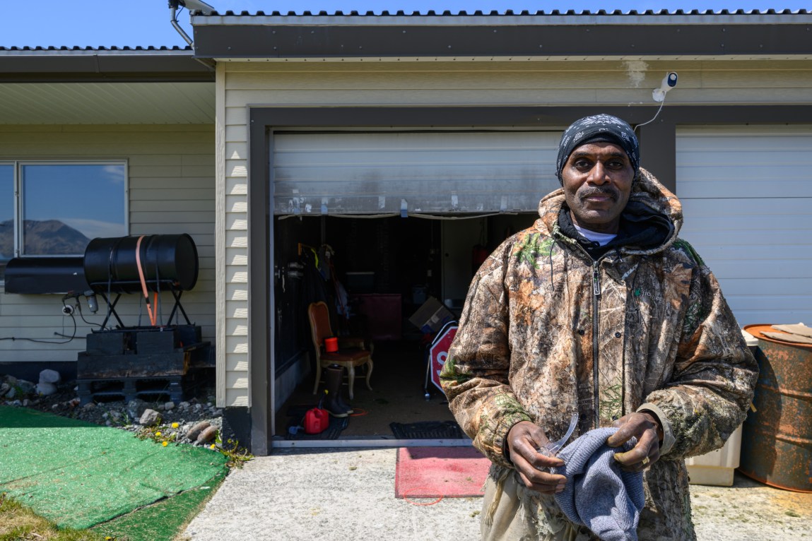 A man wearing a camo-print hooded jacket holds a gray towel and stares directly at the camera. He’s standing in front of an open garage that has a chair and a stop sign inside. A smoker sits next to the garage.