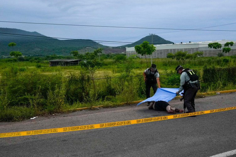 Two people in uniform drape a tarp over a dead body behind yellow police tape on the side of a road. The background is a lush green field with mountains in the distance.