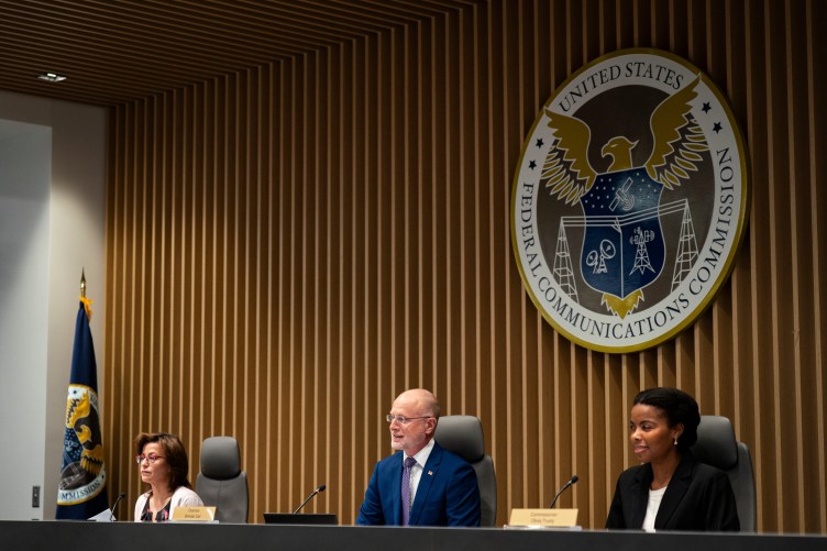 Three people, two women and a man, sit at a long desk in front of microphones. Behind them is the Federal Communications Commission logo. 
