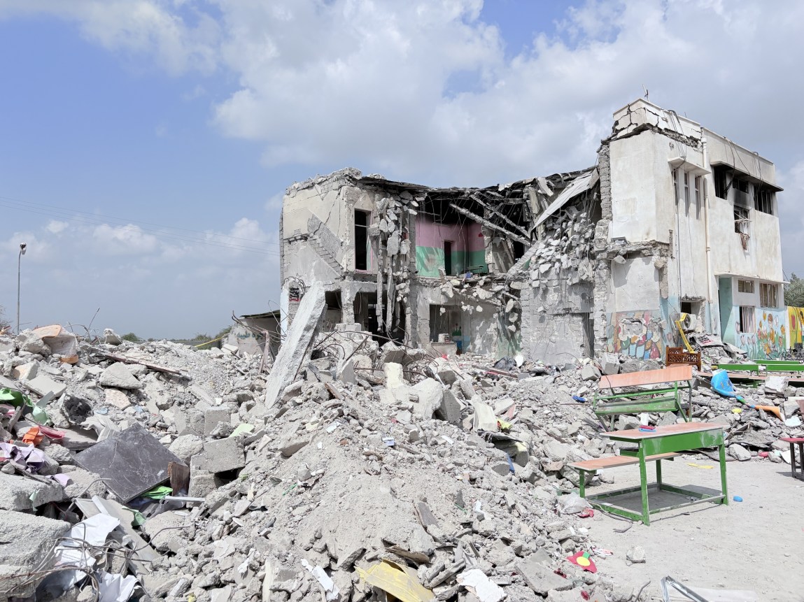 Un bâtiment en partie détruit, des tas de gravats et des pupitres d'école devant.
