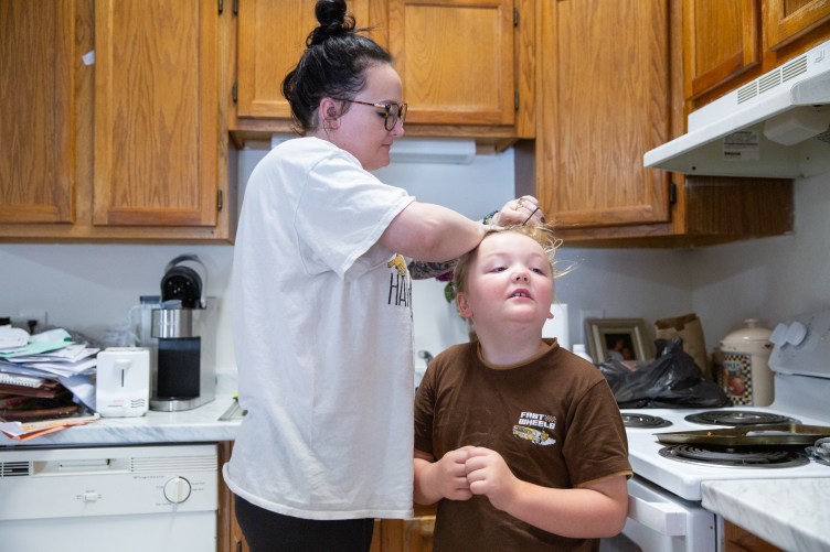 Carringer stands over a young boy wearing a brown T-shirt and puts his blond hair in a ponytail. They are standing in a kitchen in front of wooden cabinets and a stove. 