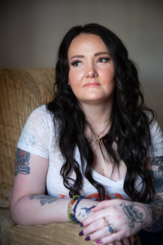 A woman with long black wavy hair sits in a brown upholstered chair and looks off camera. She wears a white V-neck T-shirt, a gold cross necklace, rings and bracelets, and she has tattoos on her arms and hands.